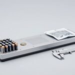 Bàn kiểm tra pin với pin trụ, pin đồng xu, túi pin và thước kẹp trên nền xám sáng — ảnh bìa bài pin và quy định pin EU